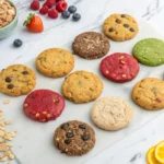 An assortment of colorful cookies laid out on a marble surface, surrounded by oats, berries, citrus slices, and nuts.