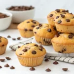 Freshly baked chocolate chip muffins on a marble plate with scattered chocolate chips and bowls in the background.