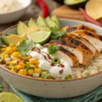A street corn chicken rice bowl with grilled chicken slices, charred corn, creamy toppings, avocado slices, and lime wedges served in a rustic ceramic bowl.