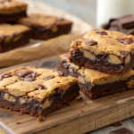 A close-up of freshly baked Brown Butter Brookies, showing the distinct layers of fudgy brownie and chewy chocolate chip cookie, stacked on a rustic wooden board.