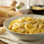 A bowl of creamy pasta garnished with black pepper and parmesan cheese, served on a wooden table with cheese, olive oil, and seasonings in the background.