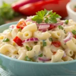 A creamy macaroni salad in a blue bowl, garnished with red onions, cherry tomatoes, and fresh parsley.