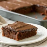 A slice of Texas sheet cake with buttermilk on a white plate, topped with glossy chocolate frosting, with the remaining cake in a baking pan in the background.